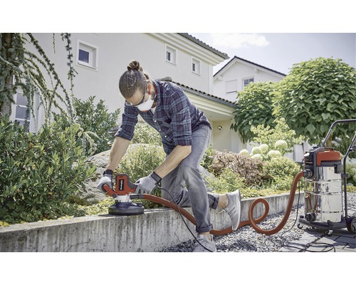 Un bărbat șlefuiește un perete de beton în grădină cu o mașină de șlefuit beton și un aspirator.