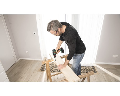 Un bărbat găurește o scândură de lemn cu o mașină de găurit cu acumulator așezată pe un banc de lucru.