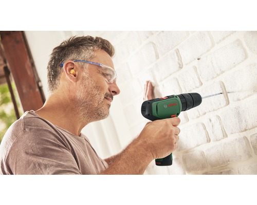 Un bărbat găurește un perete de cărămidă cu o mașină de găurit Bosch, purtând ochelari de protecție și dopuri de urechi.