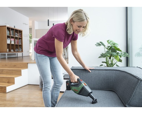 Femeie aspiră o canapea gri cu un aspirator fără fir într-un living luminos.