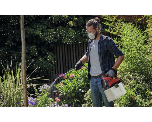 Bărbat cu ochelari de protecție și mască de respirație folosind un pulverizator de plante cu acumulator în grădină.