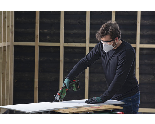 O persoană taie o placă de gips-carton cu un ferăstrău electric cu acumulator. Se poartă echipament de protecție.