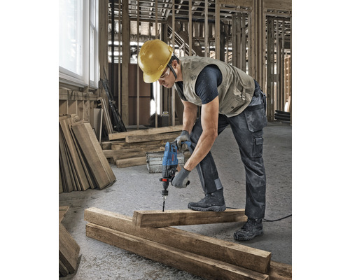 Un muncitor în construcții forează o gaură într-o scândură de lemn cu o mașină de găurit pe un șantier.