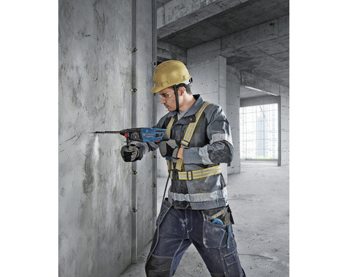 Un muncitor în construcții găurește un perete de beton cu un ciocan rotopercutor. Bărbatul poartă o cască de protecție și îmbrăcăminte de siguranță.