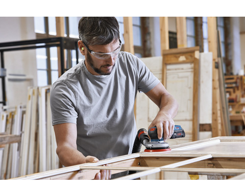 Un bărbat șlefuiește un cadru de lemn cu o șlefuitoare excentrică într-un atelier. Poartă ochelari de protecție și dopuri de urechi.