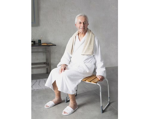 Un bărbat în halat de baie stă pe un taburet de duș cu lamele de lemn și cadru metalic.