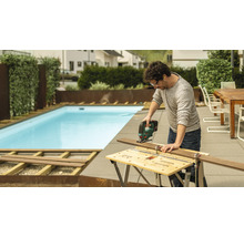 Bărbat taie o scândură de lemn cu un ferăstrău vertical în grădină, cu o piscină în fundal