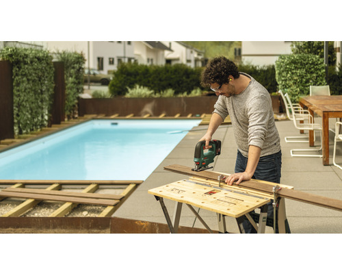 Bărbat taie o scândură de lemn cu un ferăstrău vertical în grădină, cu o piscină în fundal