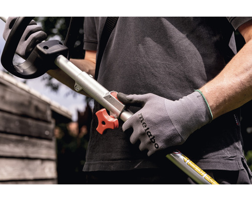 O persoană ține în mână o motocoasă cu un buton rotativ roșu și poartă mănuși de lucru.