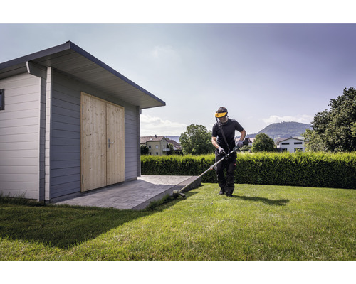 Bărbat tunde gazonul cu o mașină de tuns iarba lângă o căsuță de grădină.