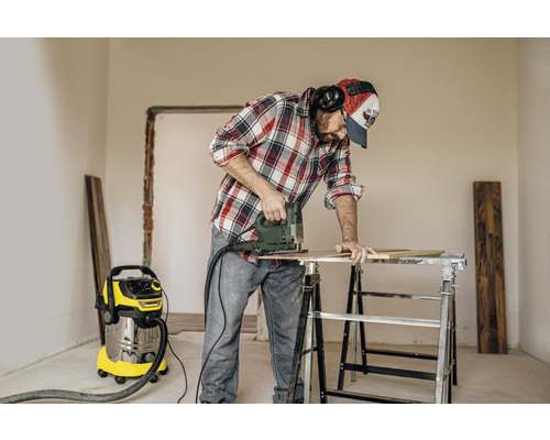 Un bărbat lucrează cu un fierăstrău vertical pe o piesă de lucru pe un banc de lucru, cu un aspirator în fundal.