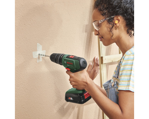 O femeie găurește un perete cu o mașină de găurit cu acumulator. Poartă ochelari de protecție.