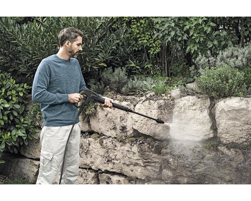 Un bărbat curăță un zid de piatră cu un aparat de curățat cu presiune înaltă în grădină.