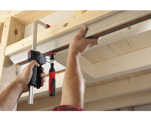 O mână fixează o șină metalică cu un clește cu șurub de o construcție din lemn.