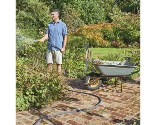 Un bărbat udă plantele cu un furtun de grădină lângă o roabă cu unelte de grădinărit.