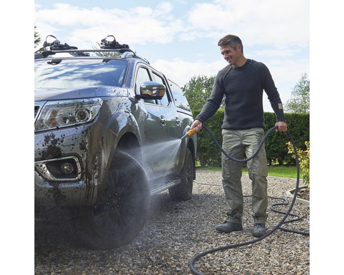 Bărbat care curăță o mașină cu un furtun de grădină pe o alee de pietriș.