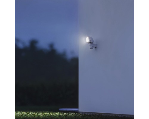 Lampa de perete exterioară cu senzor de mișcare iluminează un perete al casei pe timp de noapte.