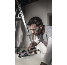 Un bărbat cu ochelari de protecție taie o piesă de lucru cu un ferăstrău circular. În fundal se află o scară.