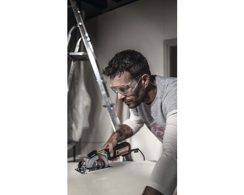 Un bărbat cu ochelari de protecție taie o piesă de lucru cu un ferăstrău circular. În fundal se află o scară.