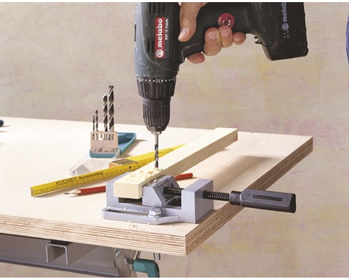 O persoană găurește cu o bormașină cu acumulator o bucată de lemn, care este fixată cu o menghină.