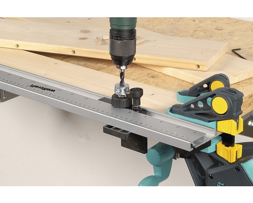 O mașină de găurit cu șablon de găurire dibluri găurește într-o scândură de lemn cu cleme pentru dibluri Wolfcraft.