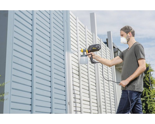 Un bărbat vopsește un gard cu un pulverizator de vopsea.