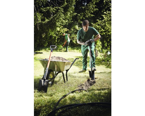 Grădinar care sapă un șanț cu o lopată lângă o roabă plină cu pământ.