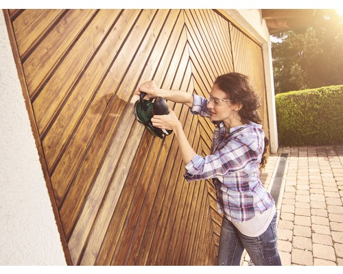 Femeie șlefuiește poarta din lemn cu un dispozitiv de șlefuit. Proiect de bricolaj în aer liber.