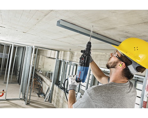 Un meșter forează cu un ciocan rotopercutor într-un tavan de beton pe un șantier.