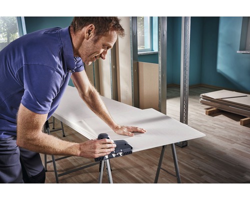 Un bărbat lucrează o placă de gips-carton cu un rindel pentru muchii pe un banc de lucru într-o construcție brută.