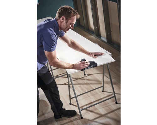 Un bărbat taie o placă de gips-carton cu o unealtă pe o masă de lucru.