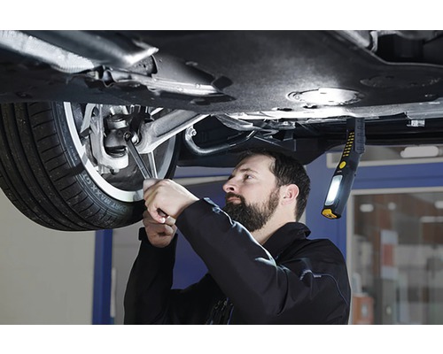 Un mecanic lucrează cu o cheie la o mașină, luminată de o lampă de lucru.