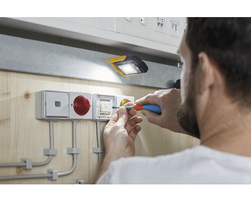 Un electrician lucrează cu o șurubelniță la o instalație într-o cameră luminoasă cu o lampă de lucru.