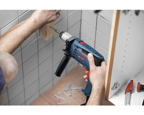 Meșterul găurește o bucată de lemn pe un perete cu o mașină de găurit cu percuție. În fundal se vede un dulap. Șuruburile sunt așezate pe podea.