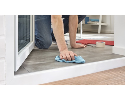 O persoană stă în genunchi și curăță o podea cu gresie cu o cârpă.