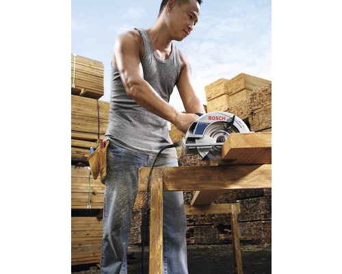 Un bărbat taie o scândură de lemn cu un ferăstrău circular, cu stive de lemn în fundal.