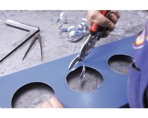 O persoană taie o gaură într-o tablă albastră cu ajutorul unei foarfeci de tablă. Un echer și un compas sunt așezate pe masă.