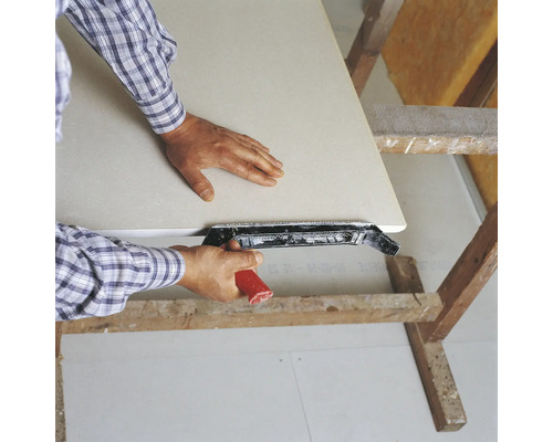 Un bărbat nivelează o placă de gips-carton cu o rindeluitoare de margini pe un capră de lemn