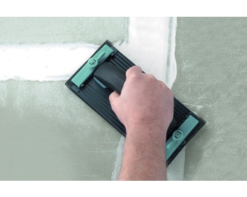 O mână folosește o placă de șlefuit pentru a netezi un perete chituit.