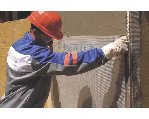 Un muncitor fixează țesătură de armare pe un perete al casei pentru izolare termică.