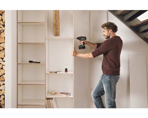 Un bărbat asamblează o bibliotecă albă cu o șurubelniță fără fir într-o cameră cu o grămadă de lemn și o scară.