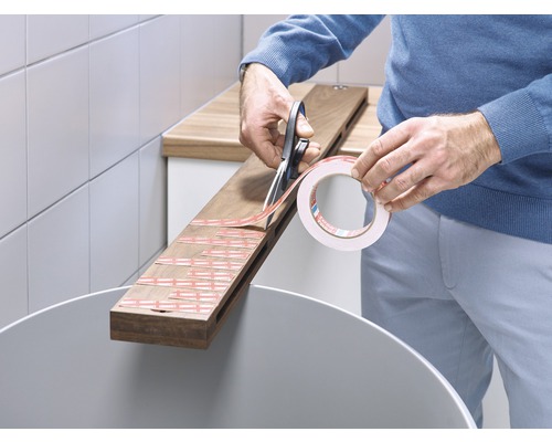 O persoană taie bandă adezivă dublă cu foarfeca pentru a o lipi pe o scândură de lemn.