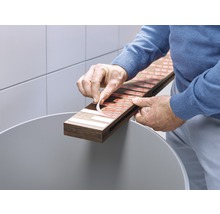 O persoană îndepărtează folii de protecție de pe benzile adezive de pe o placă de lemn ținută deasupra unei găleți.