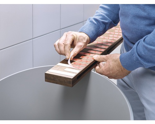 O persoană îndepărtează folii de protecție de pe benzile adezive de pe o placă de lemn ținută deasupra unei găleți.