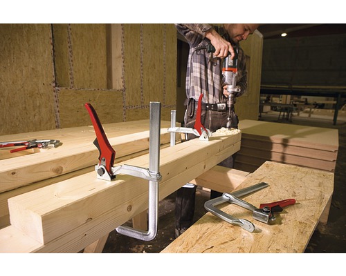 Un bărbat găurește cu o mașină de găurit o bucată de lemn fixată cu cleme de prindere