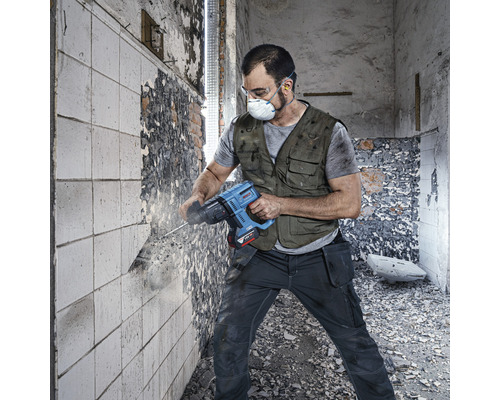 Artizan cu mască de protecție și ochelari de protecție lucrează cu un ciocan demolator pe un perete.