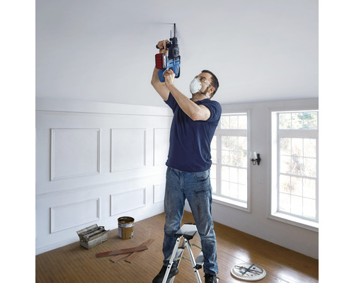 Un bărbat găurește cu o mașină de găurit o gaură în tavan stând pe o scară într-o cameră.