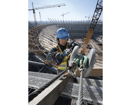 Un muncitor în construcții cu cască de protecție și vestă de siguranță lucrează cu un polizor unghiular pe un șantier.