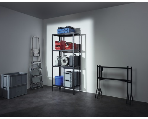 Fotografie a unui subsol cu un raft care conține cutii, cutii de scule, o rolă de cablu și o găleată, precum și o scară, două cutii pliabile gri și o masă de tapet.