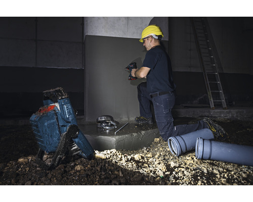 Muncitor lucrând cu o mașină de găurit, reflector de șantier și țevi de canalizare pe un șantier